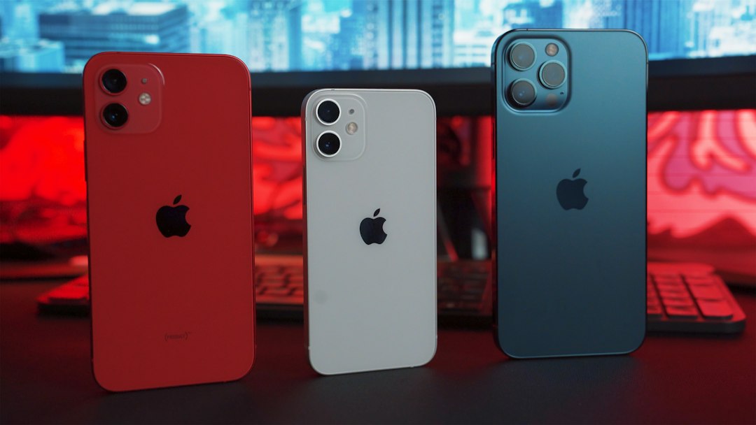 Three smartphones in red, white, and blue on a desk, representing the iPhone lineup affected by India's 2026 price hike